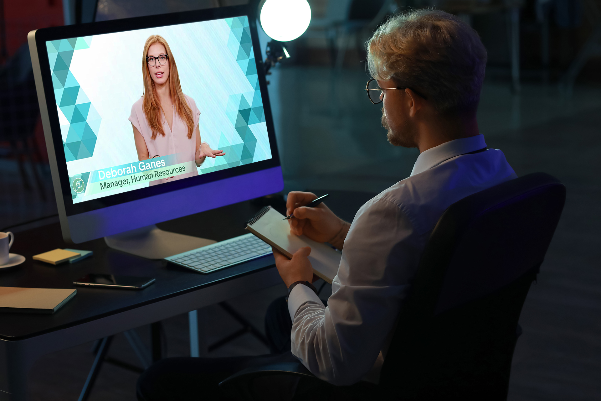 Man in office taking notes while watching a virtual meeting featuring a female presenter on screen.