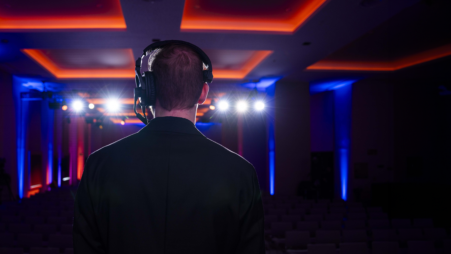 Rear view of a male Brella event management agency producer wearing headset, looking out over darkened corporate event with blue and orange accent lighting.