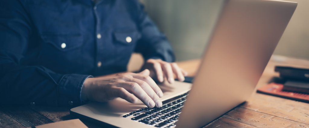 Closeup of a man in a blue shirt, typing at a laptop.