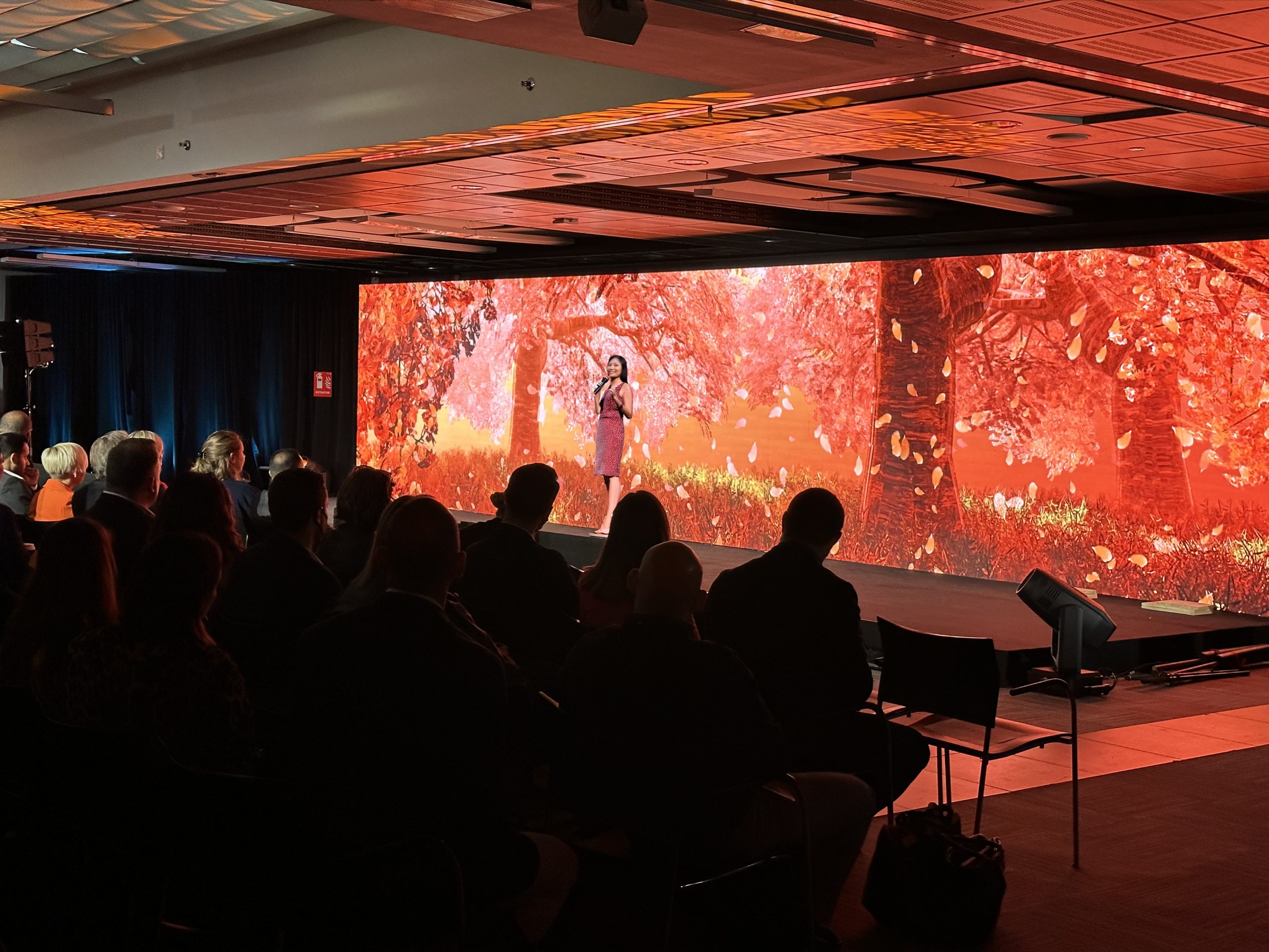 Female presenter in a dress in front of an autumnal backdrop designed by the Brella event management agency for a corporate event.