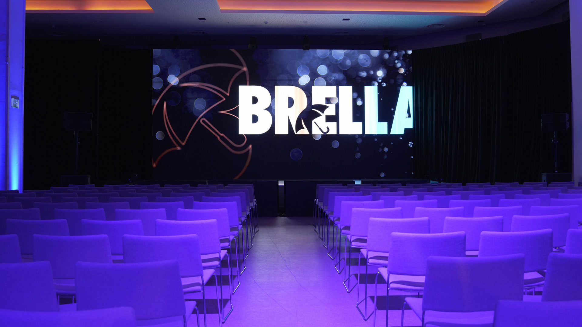 Blue lit back of house view of meeting stage with Brella logo video backdrop at corporate event produced by Brella.
