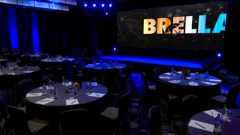 Darkened, blue lit ballroom set up for a Brella event management agency designed corporate conference session with the Brella logo on the screen.