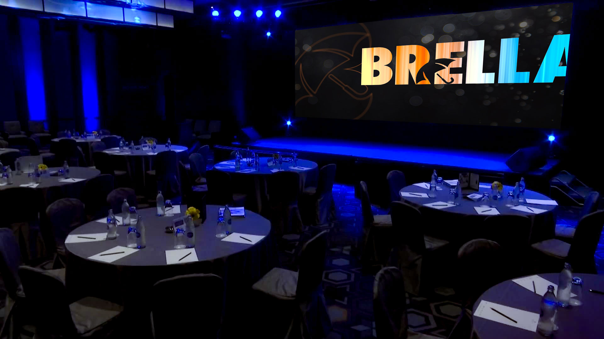 Darkened, blue lit ballroom set up for a Brella event management agency designed corporate conference session with the Brella logo on the screen.